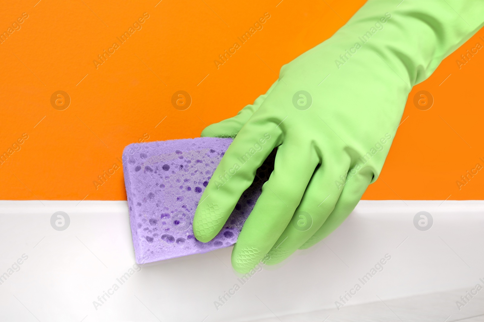 Photo of Woman in protective glove cleaning plinth with sponge indoors, closeup