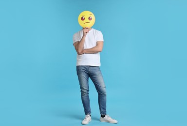 Man covering face with thinking emoticon on light blue background