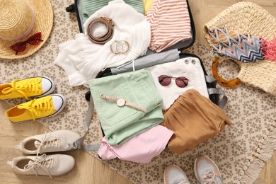 Photo of Open suitcase full of clothes and summer accessories near shoes on floor, top view