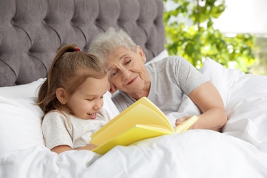 Cute girl and her grandmother reading book on bed at home