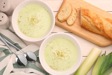 Bowls of tasty leek soup, spoons and bread on white wooden table, flat lay