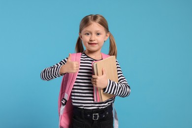 Photo of Happy schoolgirl with backpack and books showing thumb up gesture on light blue background