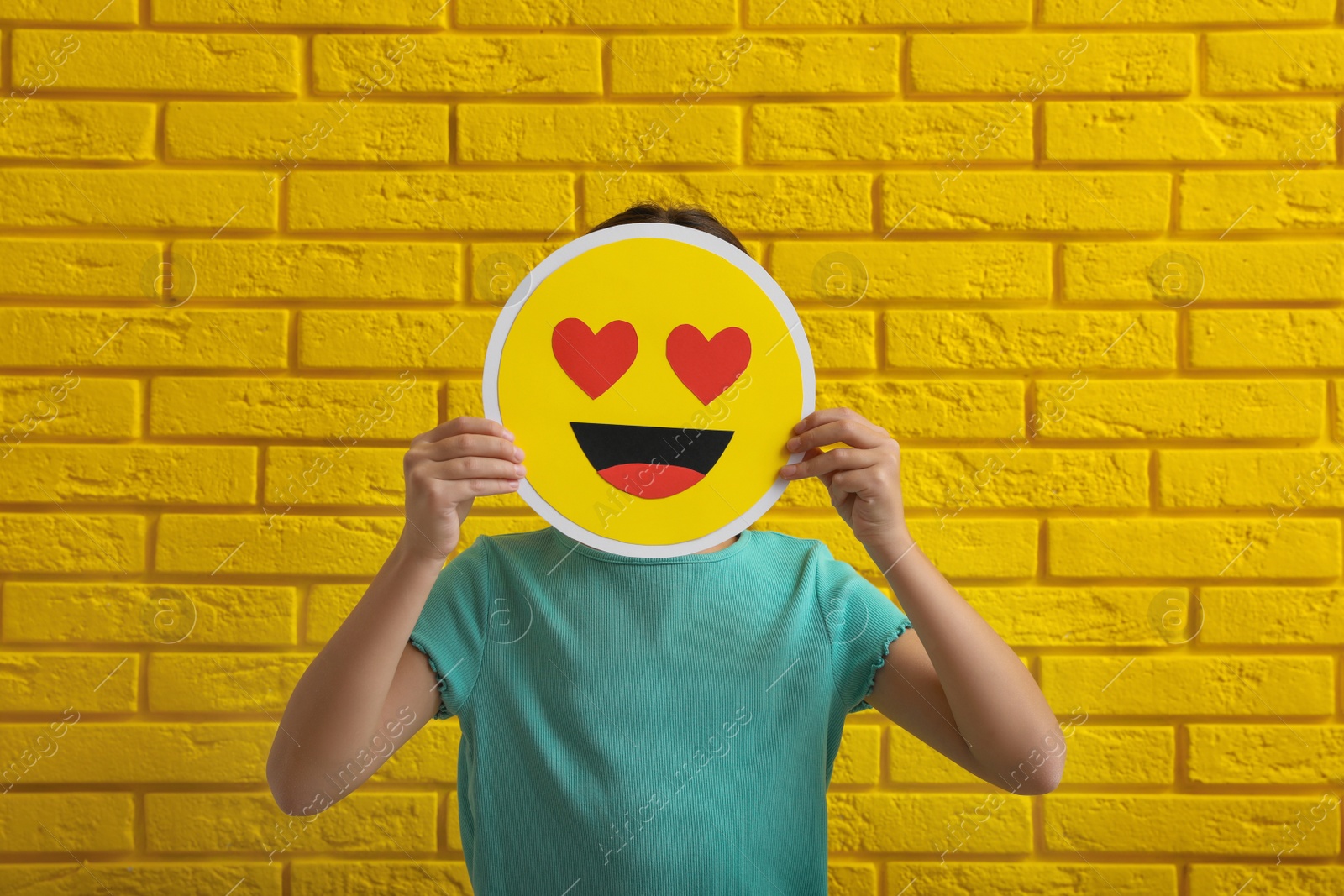 Photo of Little girl covering face with heart eyes emoji near yellow brick wall