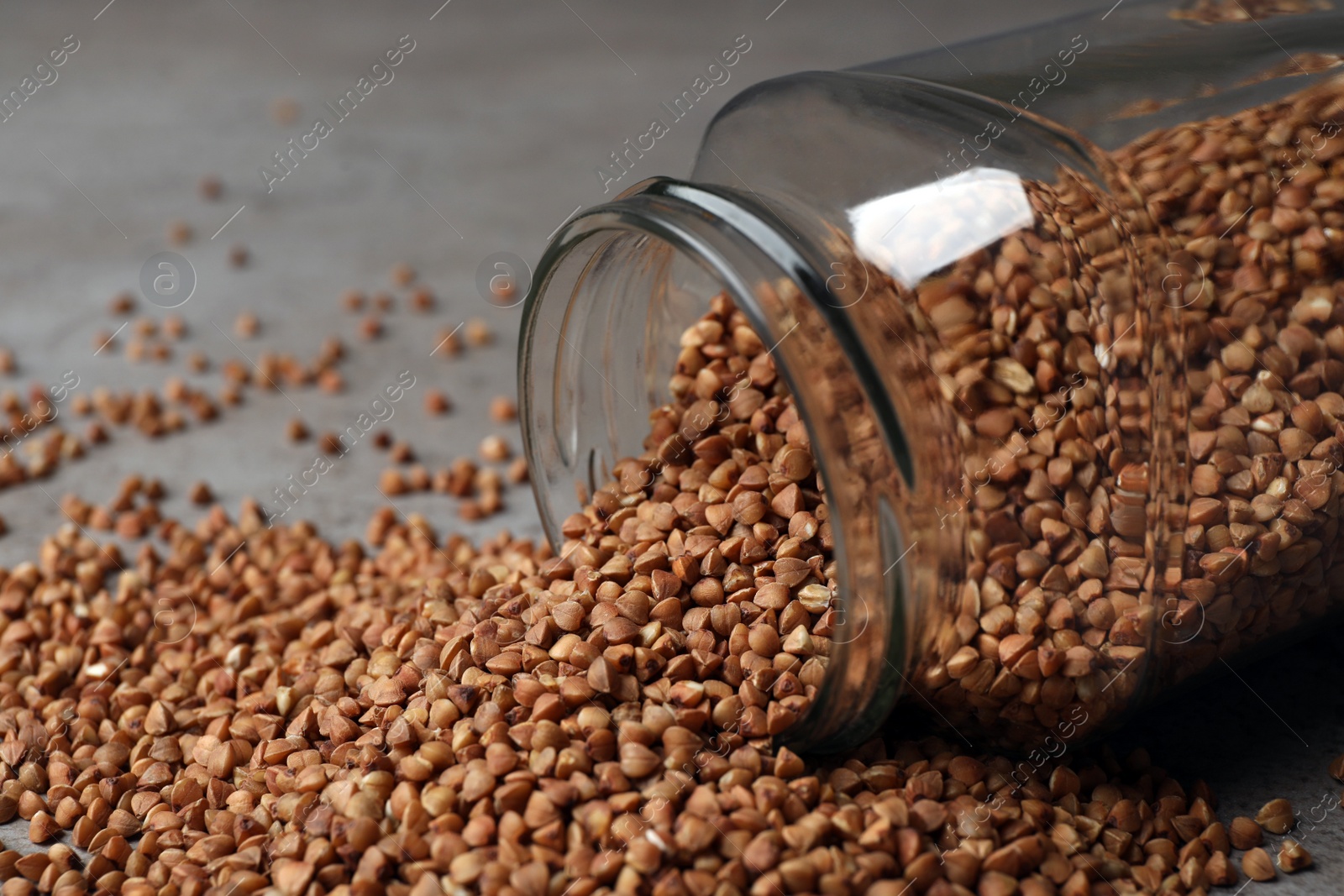 Photo of Overturned glass jar with uncooked buckwheat on table, closeup