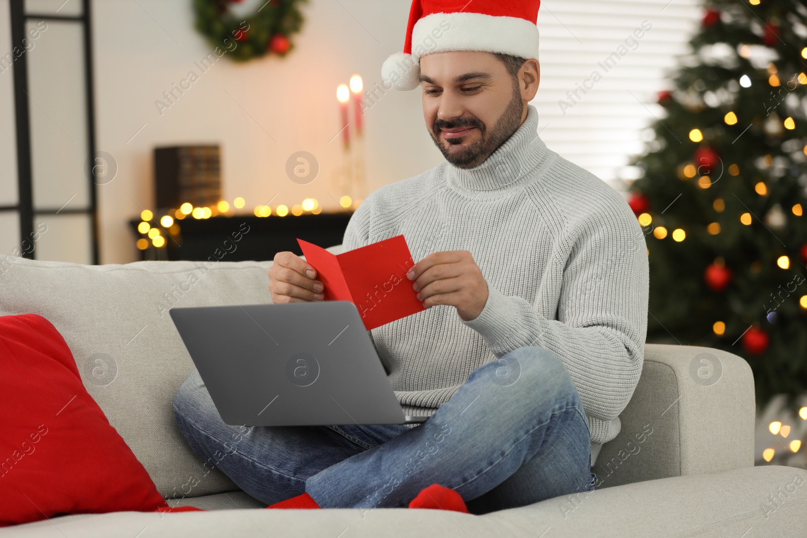 Photo of Celebrating Christmas online with exchanged by mail presents. Man in Santa hat reading greeting card during video call on laptop at home