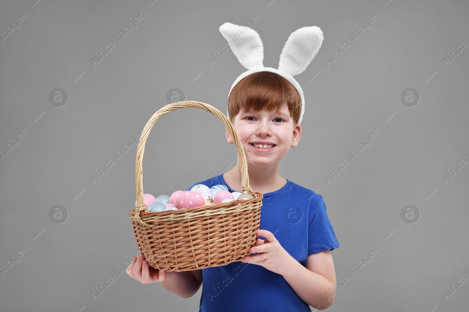 Photo of Easter celebration. Cute little boy with bunny ears and wicker basket full of painted eggs on grey background