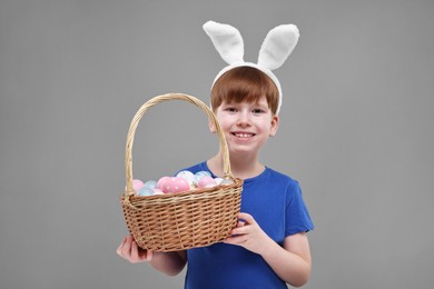 Photo of Easter celebration. Cute little boy with bunny ears and wicker basket full of painted eggs on grey background