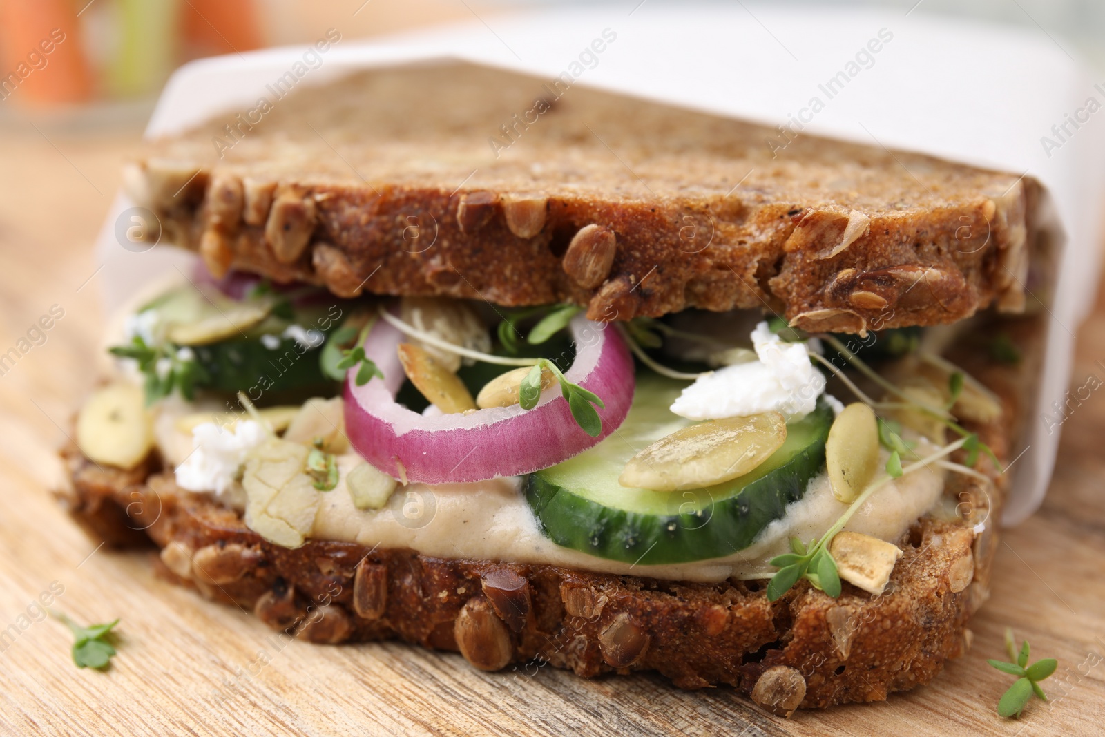 Photo of Tasty vegan sandwich with cucumber, onion, hummus and pumpkin seeds on wooden board, closeup