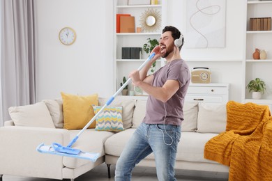 Photo of Spring cleaning. Man in headphones with mop singing while tidying up living room
