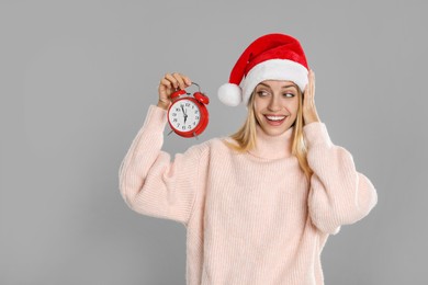 Woman in Santa hat with alarm clock on grey background. New Year countdown