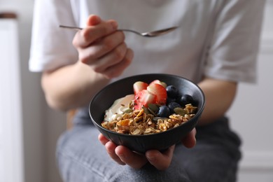 Photo of Woman eating tasty granola with berries, yogurt and seeds indoors, closeup