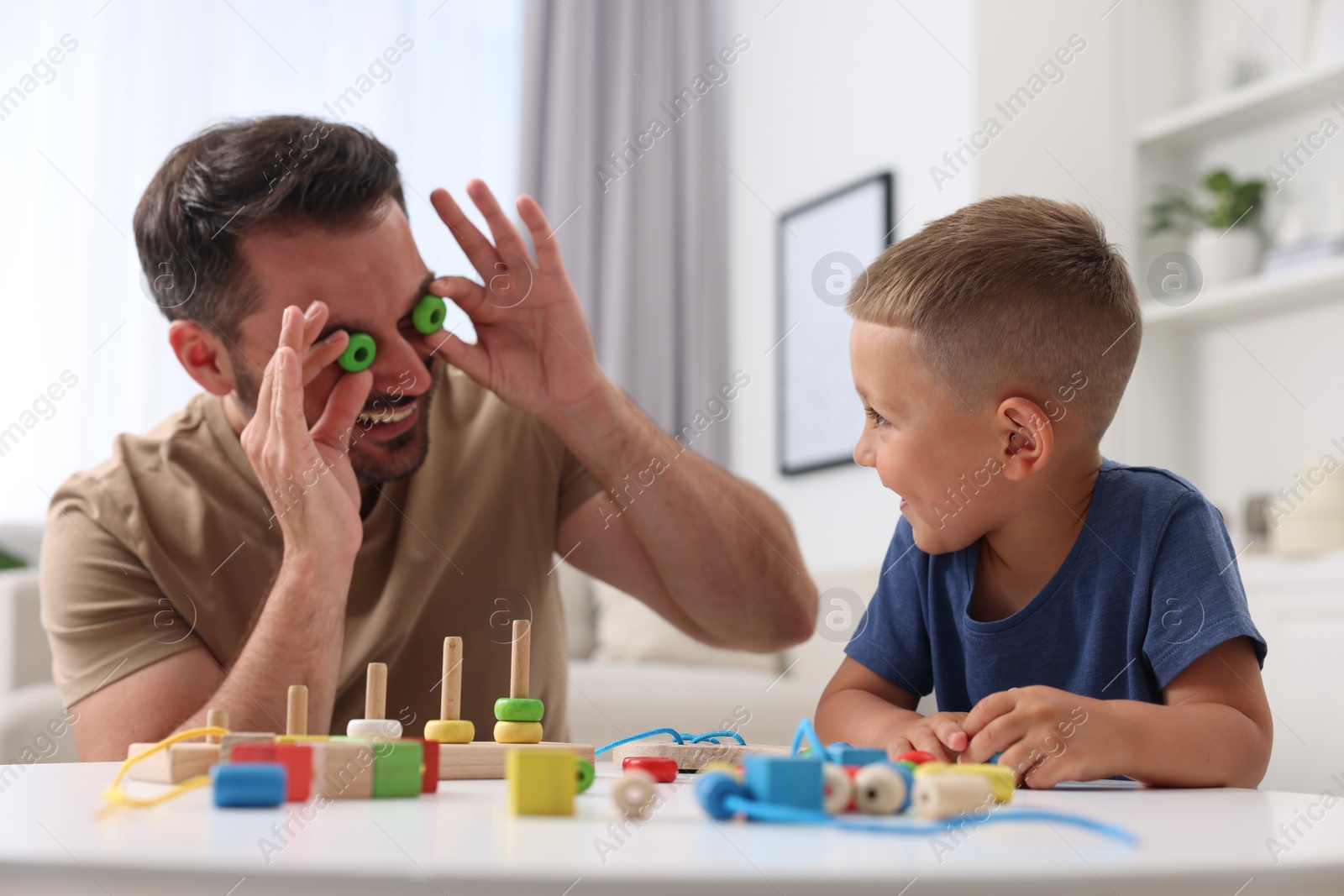 Photo of Motor skills development. Father and his son playing with wooden pieces and string for threading activity at table indoors