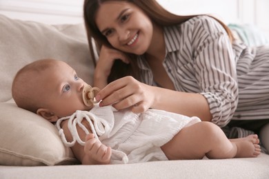 Photo of Happy mother giving pacifier to her cute little baby on sofa at home