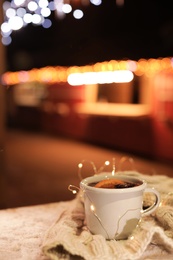 Photo of Cup of hot mulled wine and garland on table against blurred background. Space for text