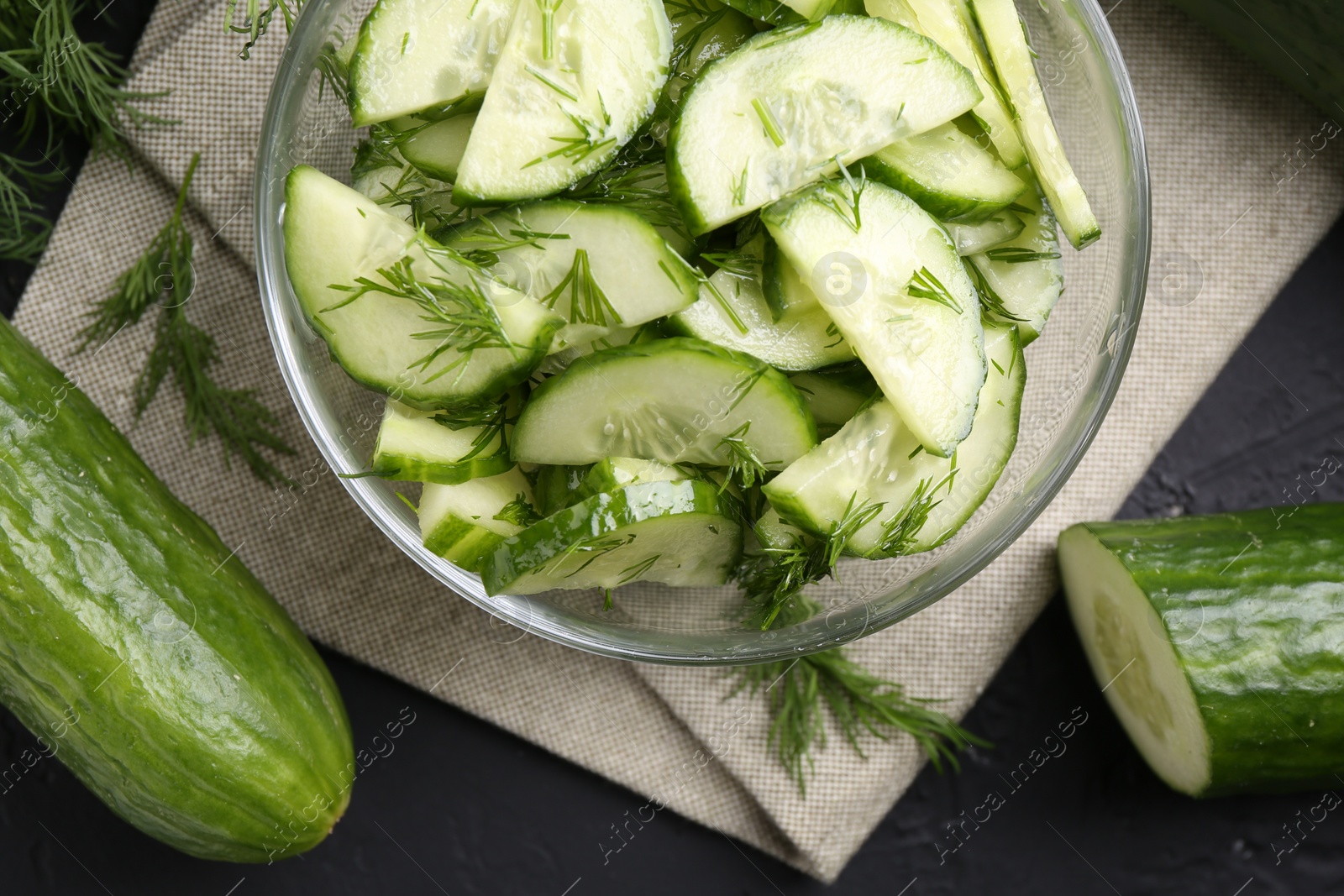 Photo of Cut cucumber with dill in glass bowl and fresh vegetables on dark gray table, flat lay