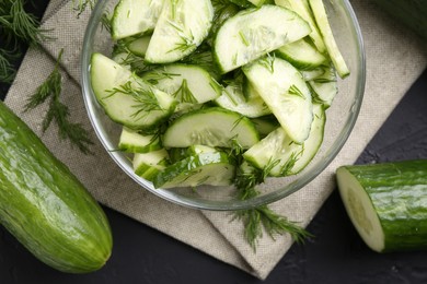 Photo of Cut cucumber with dill in glass bowl and fresh vegetables on dark gray table, flat lay