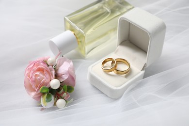 Wedding stuff. Stylish boutonniere, wedding rings and perfume bottle on white veil, closeup