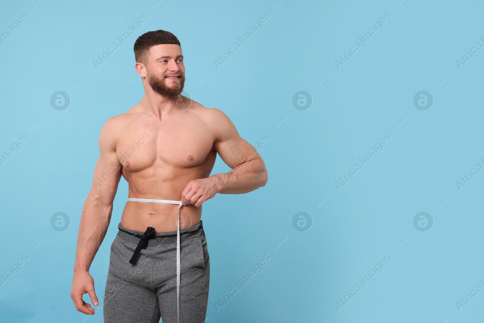 Photo of Portrait of happy athletic man measuring waist with tape on light blue background, space for text. Weight loss concept