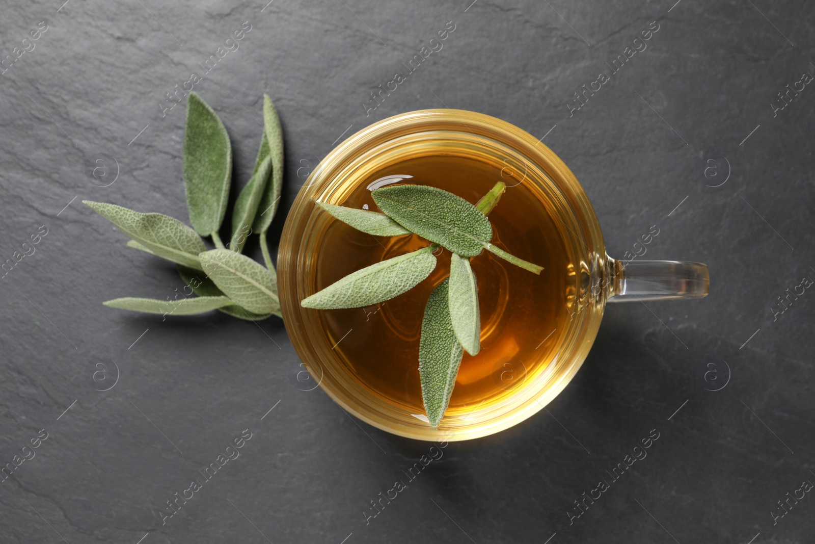 Photo of Cup of aromatic sage tea and fresh leaves on black table, flat lay