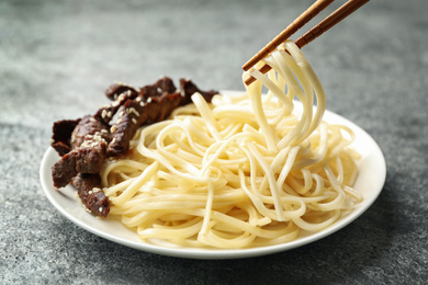 Chopsticks with tasty cooked rice noodles over plate on grey table