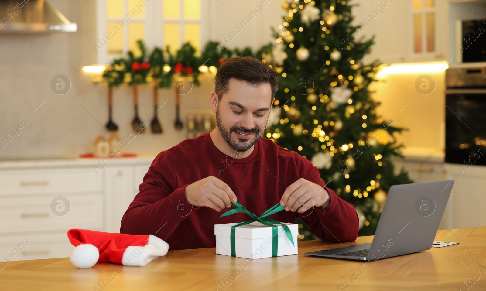 Photo of Celebrating Christmas online with exchanged by mail presents. Man opening gift box during video call on laptop at home