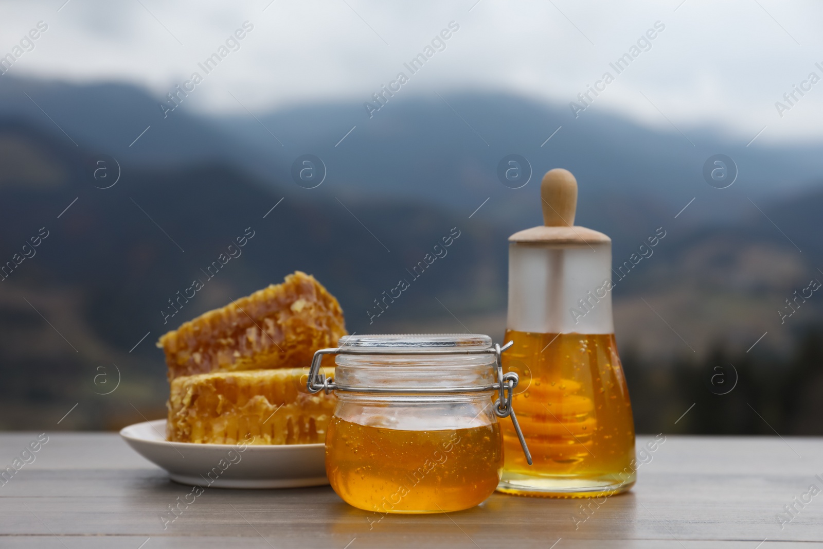 Photo of Fresh aromatic honey and combs on grey wooden table against mountain landscape