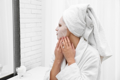 Photo of Beautiful woman with sheet mask on her face at mirror indoors