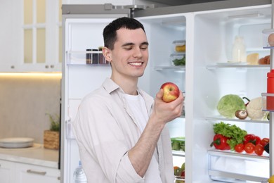 Happy man holding apple near refrigerator in kitchen