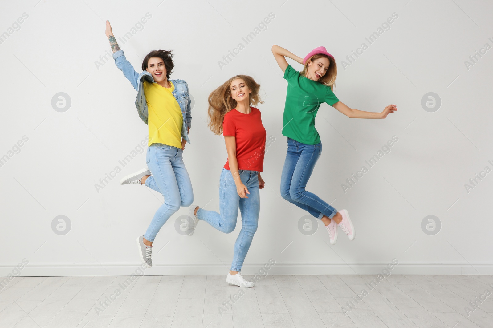 Photo of Group of young women in jeans and colorful t-shirts jumping near light wall