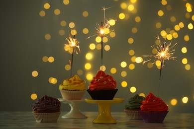 Photo of Different colorful cupcakes with sparklers on table against blurred lights