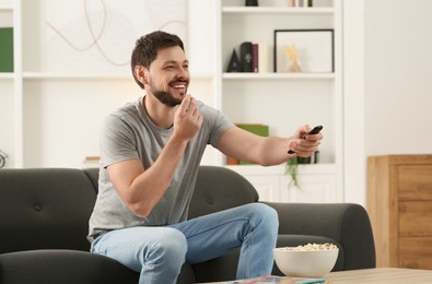 Photo of Happy man eating popcorn while changing TV channels with remote control on sofa at home