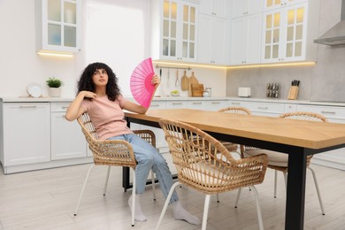 Photo of Young woman waving pink hand fan to cool herself at table in kitchen