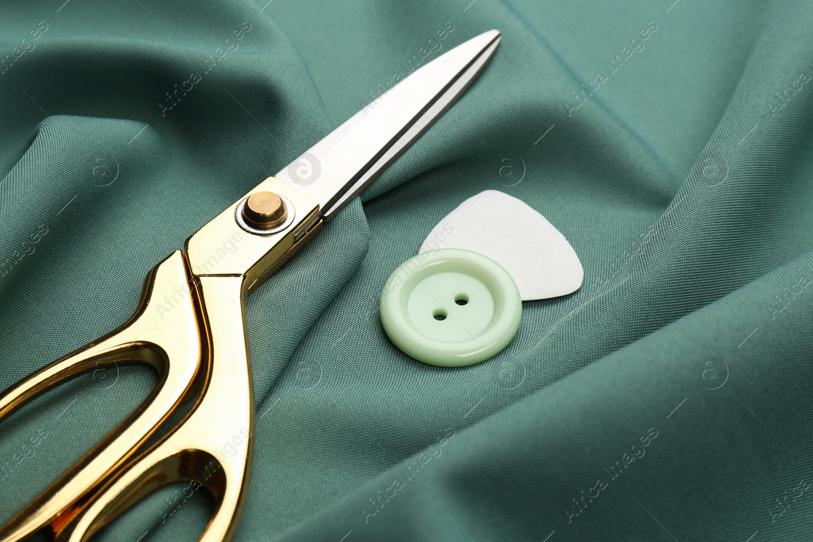 Photo of Scissors, button and tailor's chalk on green fabric, closeup
