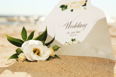 Photo of Envelope with wedding invitation and beautiful flower on sandy beach, closeup