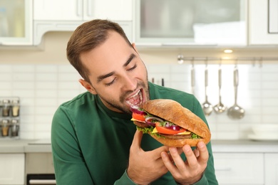 Young hungry man eating tasty sandwich in kitchen