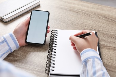 Photo of Woman with smartphone writing in notebook at wooden table, closeup