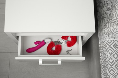 Photo of Pink vibrator, fluffy handcuffs and keys in open drawer of bedside table indoors, above view. Sex toys