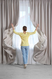 Woman opening window curtains at home in morning, back view