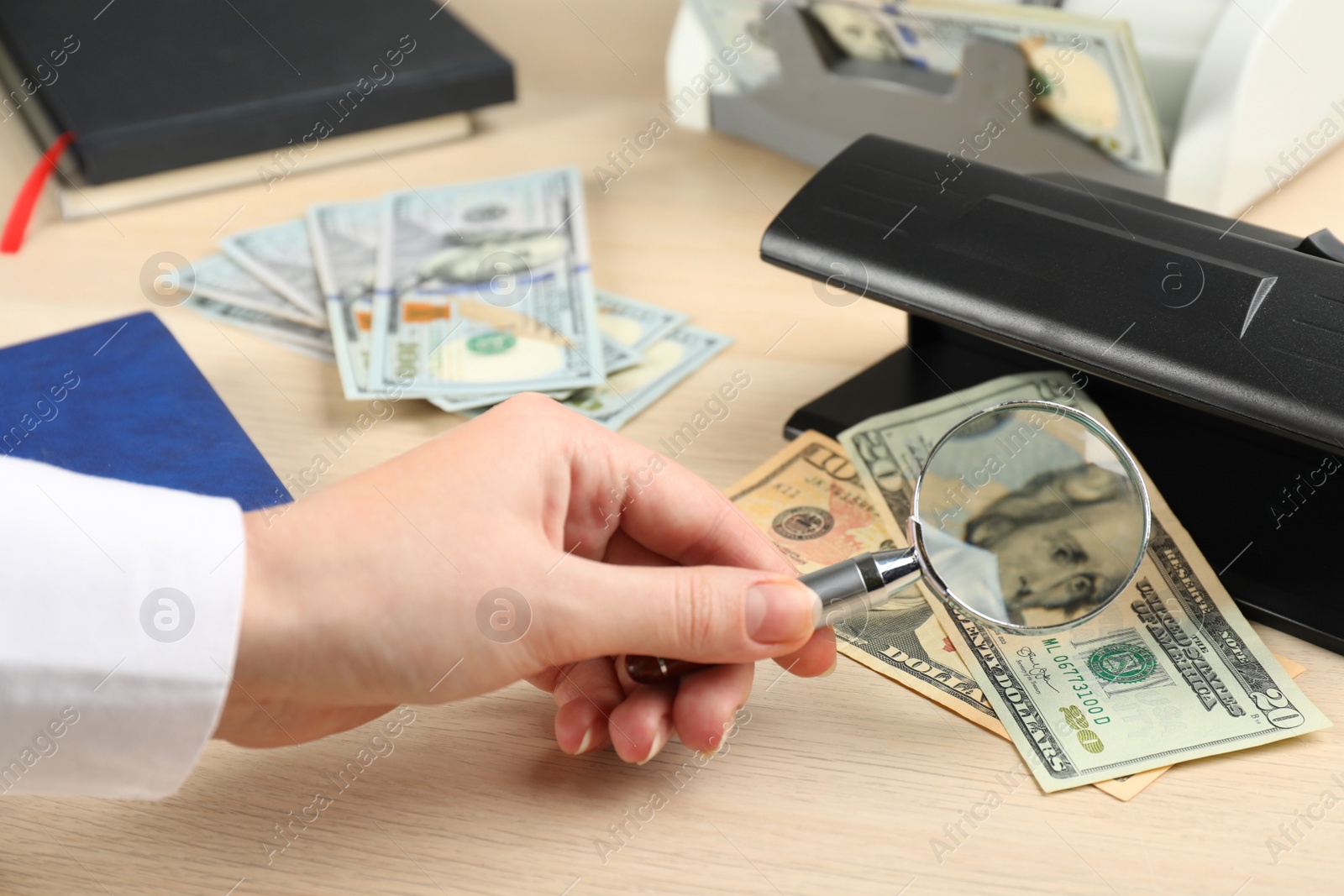 Photo of Woman checking dollar banknotes with currency detector and magnifying glass at wooden table, closeup. Money examination device