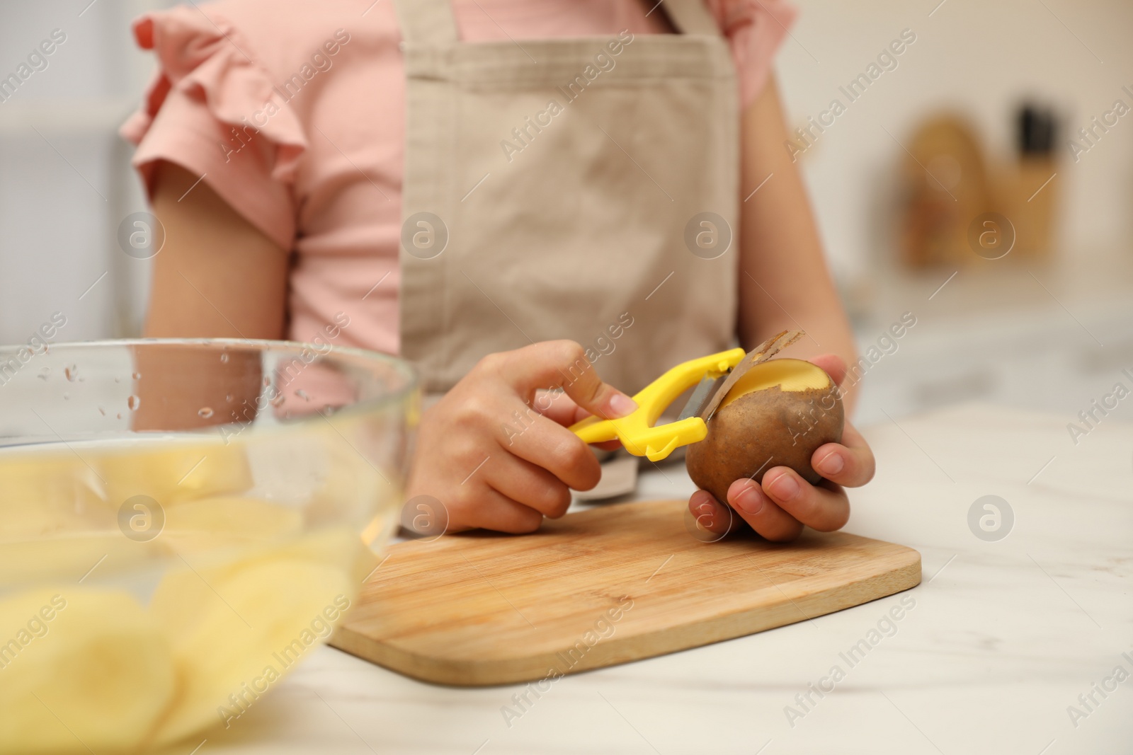 Photo of Little girl peeling potato at table in kitchen, closeup. Preparing vegetable