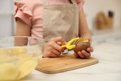 Little girl peeling potato at table in kitchen, closeup. Preparing vegetable