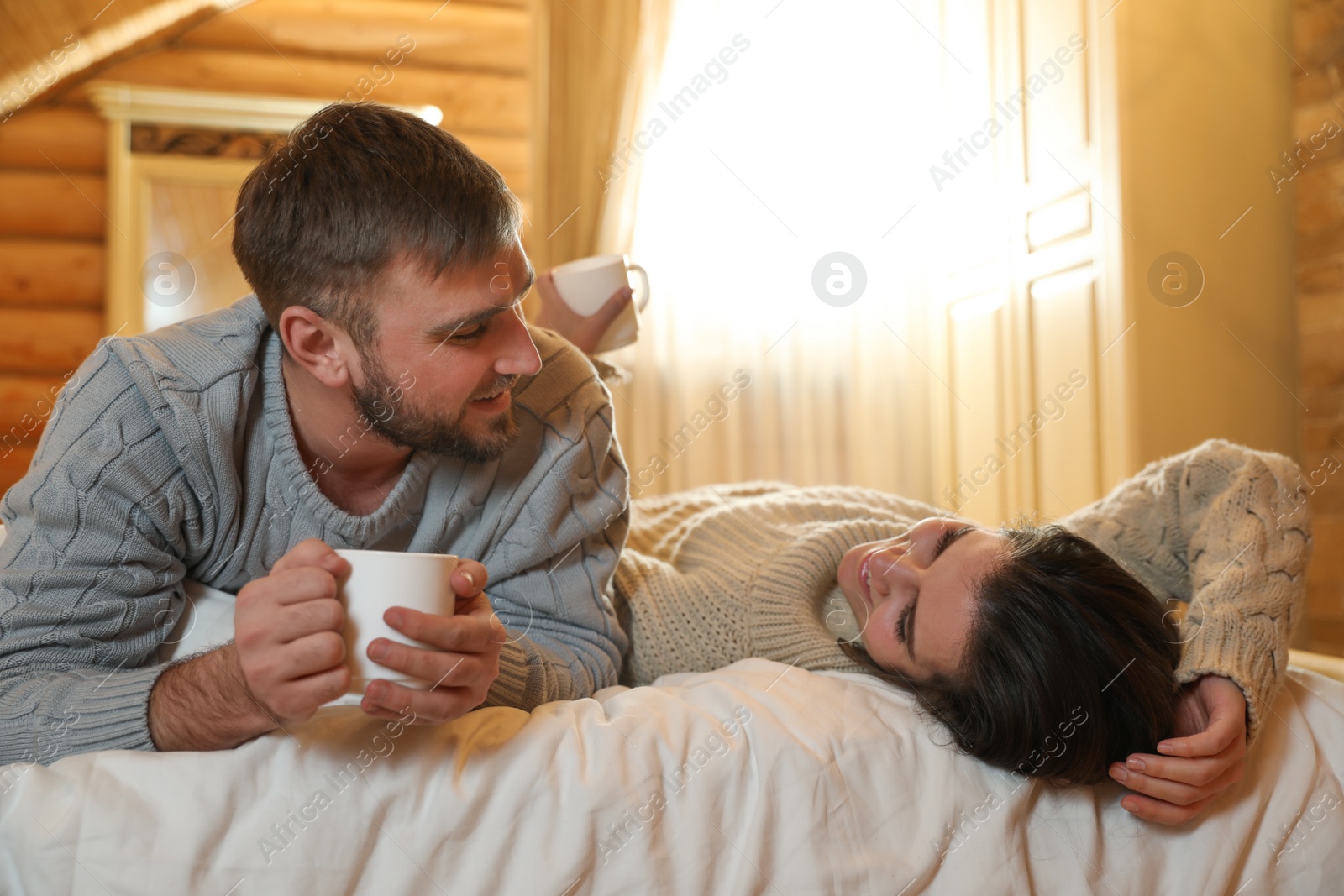 Photo of Young couple in warm sweaters with cups of hot drink on bed at home