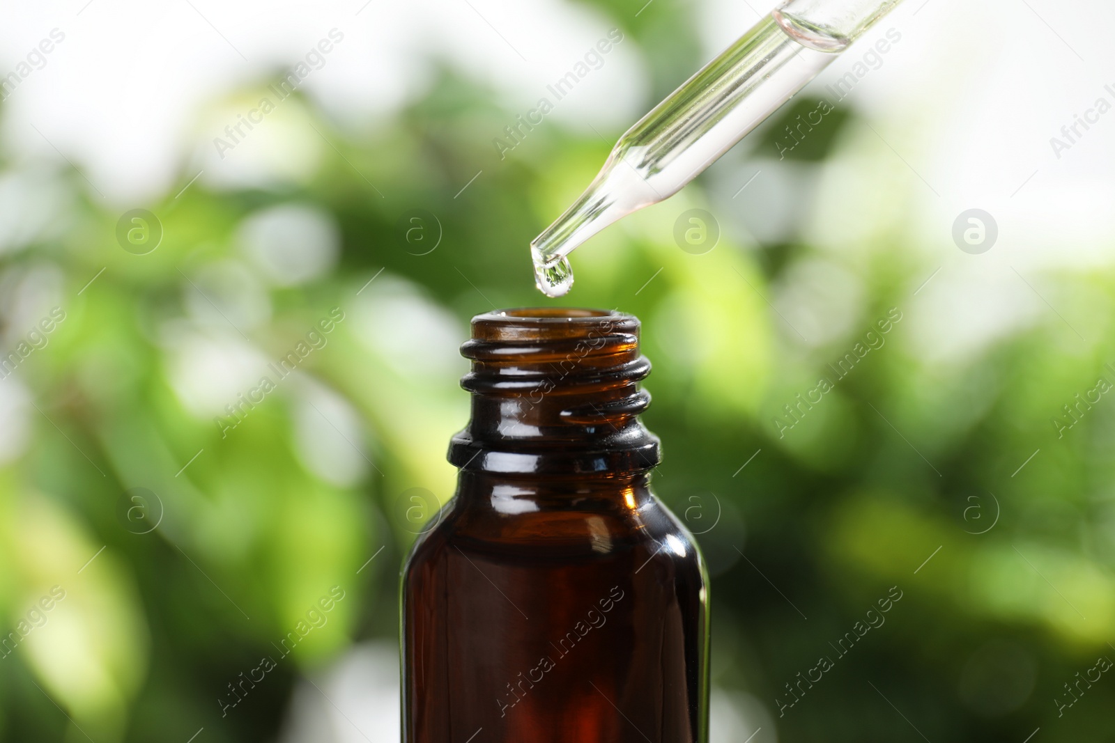 Photo of Dropper with essential oil over bottle against blurred background, closeup