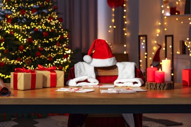 Photo of Santa's Claus workplace. Gift boxes, letters, burning candles on table and costume in room with Christmas decor