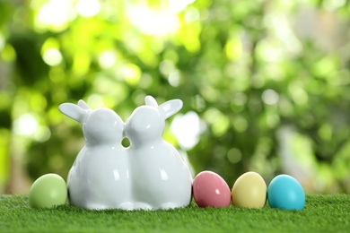 Cute ceramic Easter bunnies and dyed eggs on green grass against blurred background, space for text