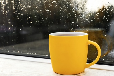 Yellow cup of hot tea on white wooden window sill, closeup. Rainy weather