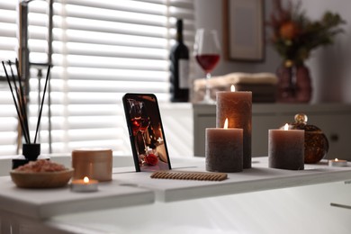 White wooden tray with smartphone, burning candles and beauty products on bathtub in bathroom
