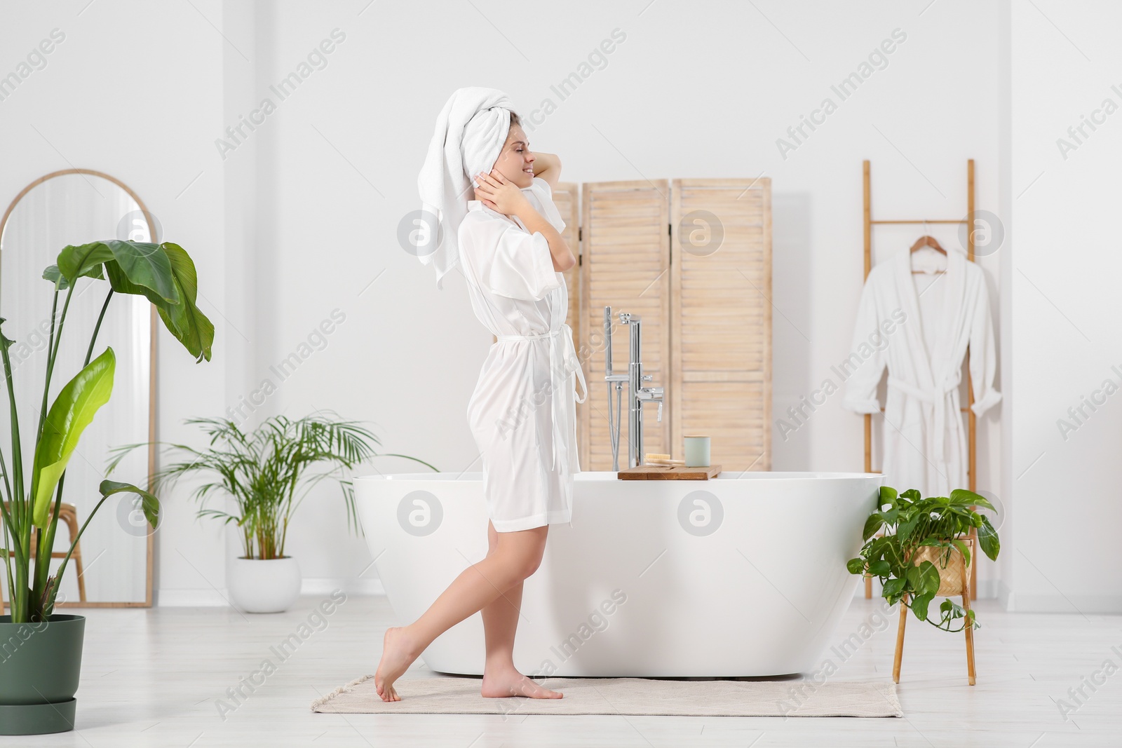 Photo of Beautiful happy woman wearing white robe near tub in bathroom