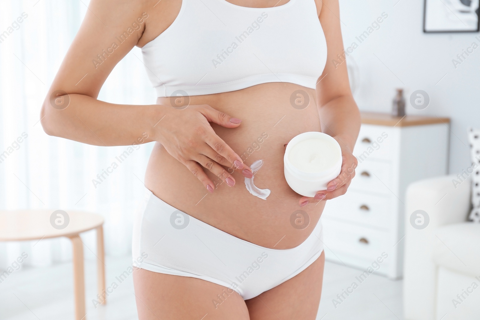 Photo of Pregnant woman applying body cream on belly at home, closeup