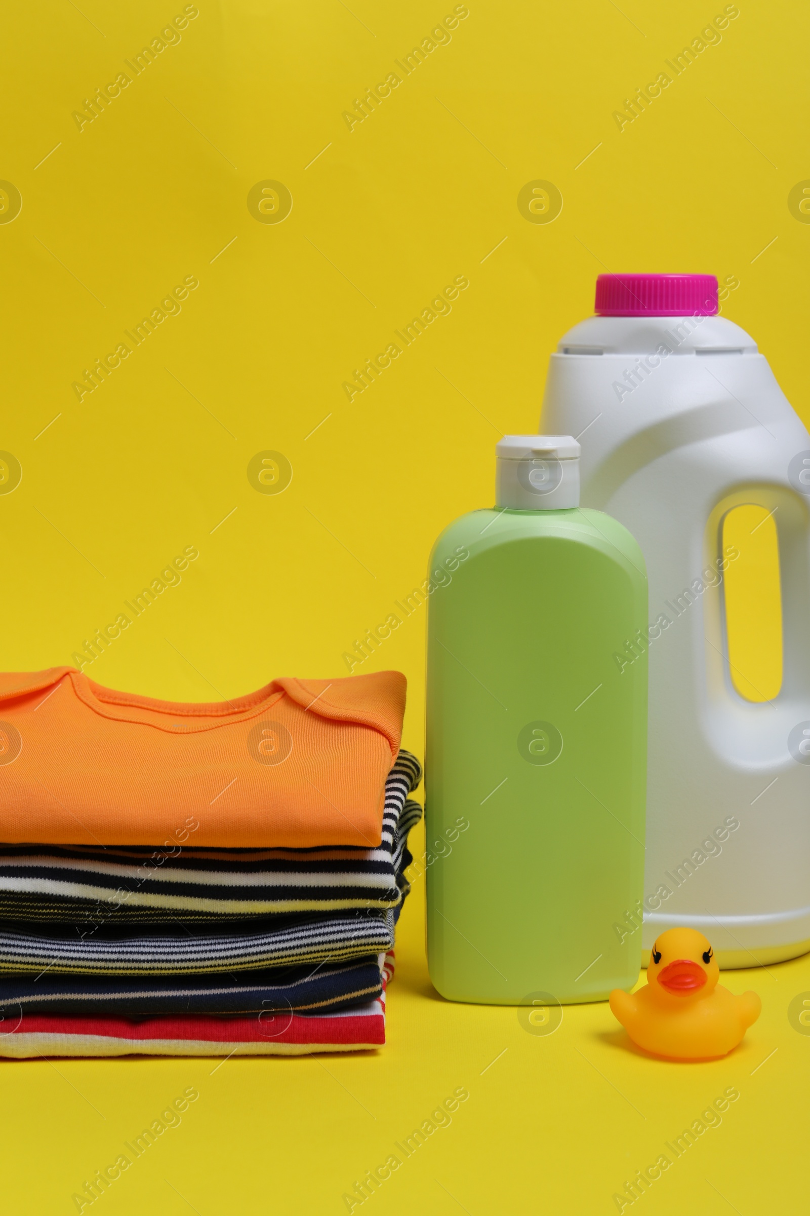 Photo of Laundry detergents, stack of baby clothes and rubber duck on yellow background